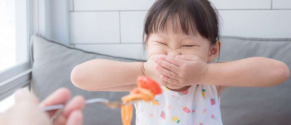 A child covers her mouth with both hands while being approached with a fork with food on it.